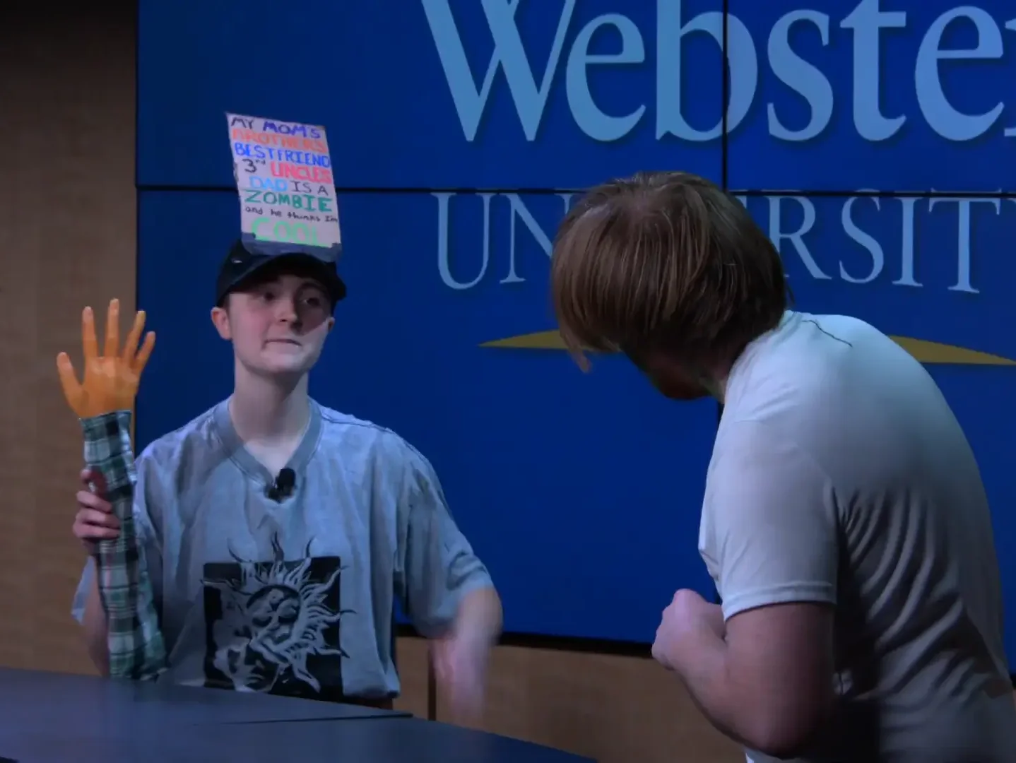 A young woman with a sign on her head holds up an arm to a zombie